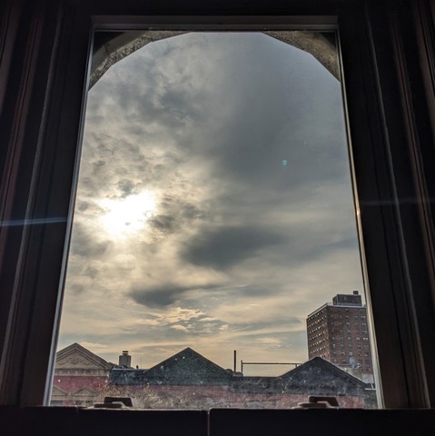 Looking through an arched window two hours after sunrise the sky is full of murky light gray clouds and the sun is breaking through on the left. Pointed roofs of Harlem brownstones with red brickwork are across the street, and a taller apartment building can be seen in the distance. The window is grimy on the bottom.