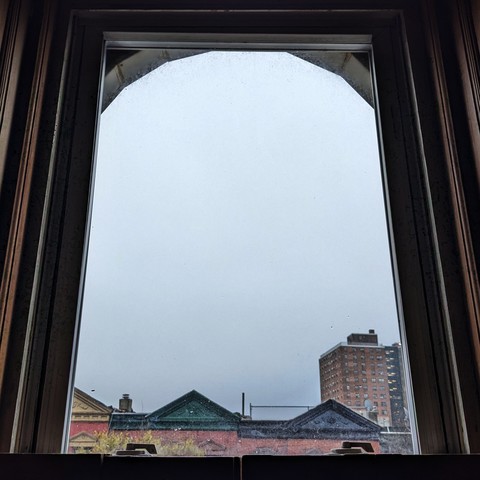 Looking through an arched window an hour after sunrise the sky is full of murky light gray clouds. Pointed roofs of Harlem brownstones with red brickwork are across the street, and a taller apartment building can be seen in the distance. The green shoots of a tree are entering the frame on the bottom left.