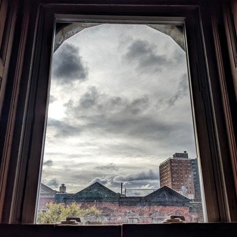 Looking through an arched window two hours after sunrise the sky is full of scattered gray clouds. Pointed roofs of Harlem brownstones with red brickwork are across the street, and a taller apartment building can be seen in the distance. The window is grimy on the bottom. The green shoots of a tree are entering the frame on the bottom left.