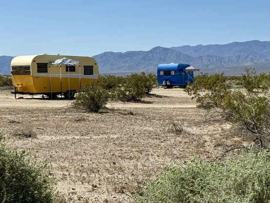 Wide iPhone shot of two cute little travel trailers, 1 bright yellow, one bright blue, sitting out in the desert; there’s lots of sandy foreground with desert scrub & in the background are dark blue mountains & a bright blue summer sky