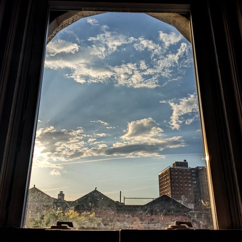 Looking through an arched window an hour after sunrise fluffy white, windswept clouds are passing through the blue sky, illuminated by the sun which is just out of frame on the left. Pointed roofs of Harlem brownstones with red brickwork are silhouetted across the street, and a taller apartment building can be seen in the distance. The window is grimy on the bottom. The green shoots of a tree are entering the frame on the bottom left.