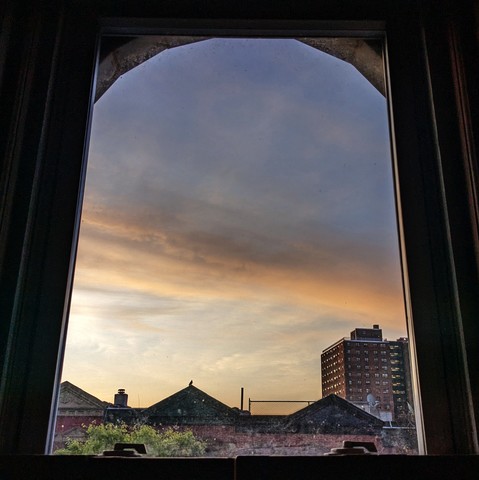 Looking through an arched window at the moment of sunrise the pale blue sky is bisected by a dramatic pale yellow cloud. Pointed roofs of Harlem brownstones with red brickwork are silhouetted across the street, and a taller apartment building can be seen in the distance. The window is grimy on the bottom. The green shoots of a tree are entering the frame on the bottom left.