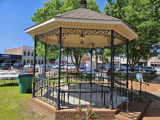 A gazebo with black wrought iron supports and fencing with a white roof in a park square.  One and two story buildings with shops in the background.