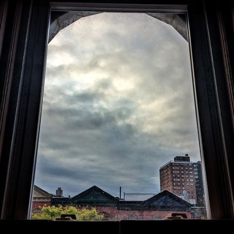 Looking through an arched window forty six minutes after sunrise the sky is full of complex blue-grey clouds. Pointed roofs of Harlem brownstones with red brickwork are across the street, and a taller apartment building can be seen in the distance. The green shoots of a tree are entering the frame on the bottom left.
