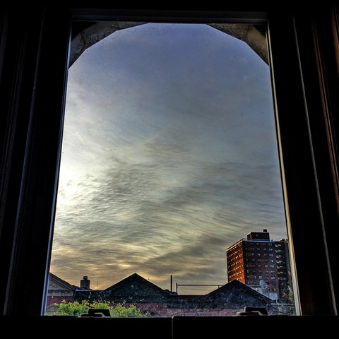 Looking through an arched window eleven minutes after sunrise the blue sky is marked with small wavy grey clouds and a peach haze is drifting from the left. Pointed roofs of Harlem brownstones with red brickwork are across the street, and a taller apartment building can be seen in the distance. The green shoots of a tree are entering the frame on the bottom left.