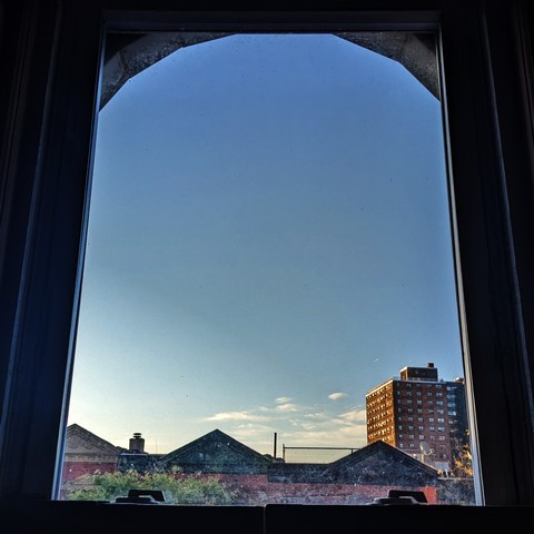 Looking through an arched window forty-five minutes after sunrise the blue sky is marked with a few small white small clouds just above the skyline. Pointed roofs of Harlem brownstones with red brickwork are across the street, and a taller apartment building can be seen in the distance. The green shoots of a tree are entering the frame on the bottom left.