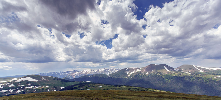 Panoramic photo of the Never Summer Range of mountains in Rocky Mountain National Park, viewed from Trail Ridge Road. In the foreground there is some reddish-brown tundra vegetation, behind which is an expanse of rolling terrain with patches of snow. Across the middle of the frame is the range of mountains with lots of snow above the tree line. Above everything there are white cumulus clouds with spots of blue sky. July 2019.