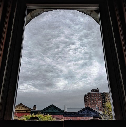 Looking through an arched window forty-nine minutes after sunrise the sky is full of dense swirling gray clouds. Pointed roofs of Harlem brownstones with red brickwork are across the street, and a taller apartment building can be seen in the distance. The window is grimy on the bottom. The green leaves of trees are entering frame on the bottom left and right.