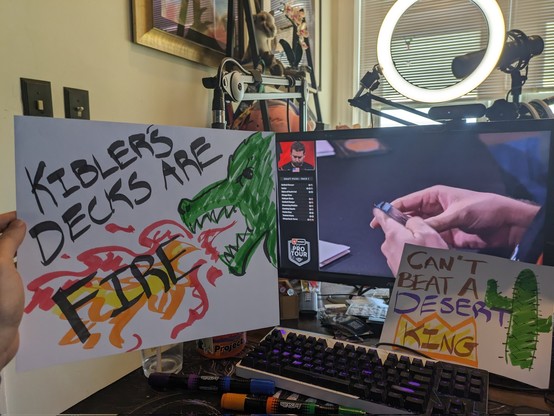 A picture of Amy's streaming setup in the background. In the foreground are 2 more signs. One says "Kibbler's decks are FIRE". The other says "Can't beat a Desert KING"