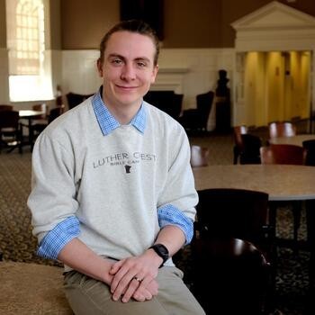 A man smiling at the camera, sitting on the edge of a table, his hands crossed on his lap. He wears a heather-grey sweatshirt with the words Luther Crest Bible Camp on it. The collar and cuffs of a blue checked button up shirt are visible.