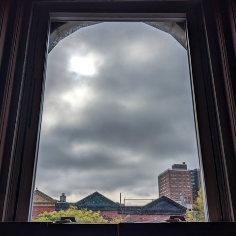 Looking through an arched window three hours after sunrise the sky is full of lumpy gray clouds. Pointed roofs of Harlem brownstones with red brickwork are across the street, and a taller apartment building can be seen in the distance. The green leaves of trees are entering frame on the bottom left and right.