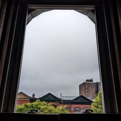Looking through an arched window two hours after sunrise the sky is full of gray clouds. Pointed roofs of Harlem brownstones with red brickwork are across the street, and a taller apartment building can be seen in the distance. The green leaves of trees are entering frame on the bottom left and right.