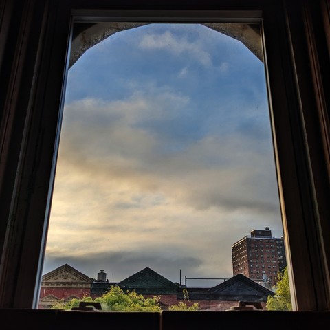 Looking through an arched window half an hour after sunrise the blue sky is smeared with parchment and gray clouds. Pointed roofs of Harlem brownstones with red brickwork are across the street, and a taller apartment building can be seen in the distance. The green leaves of trees are entering frame on the bottom left and right.