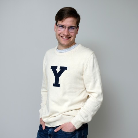 A man wearing glasses, smiles at the camera while wearing a cream colored collegiate sweater with the Yale letter "Y" on the front in Yale blue. His hands are in the pockets of his blue jeans.