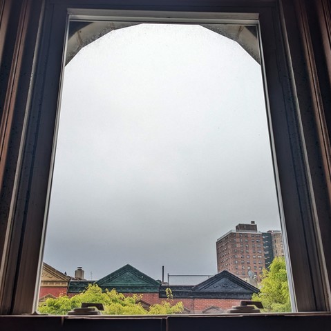 Looking through an arched window one hundred minutes after sunrise the sky is full of fog, fading from slate grey at the horizon to almost white up above. Pointed roofs of Harlem brownstones with red brickwork are  across the street, and a taller apartment building can be seen in the distance. The green leaves of trees are entering frame on the bottom left and right.