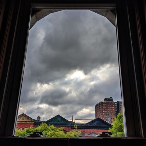 Looking through an arched window ninety minutes after sunrise the sky is full of puffy gray clouds. Pointed roofs of Harlem brownstones with red brickwork are across the street, and a taller apartment building can be seen in the distance. The green leaves of trees are entering frame on the bottom left and right.