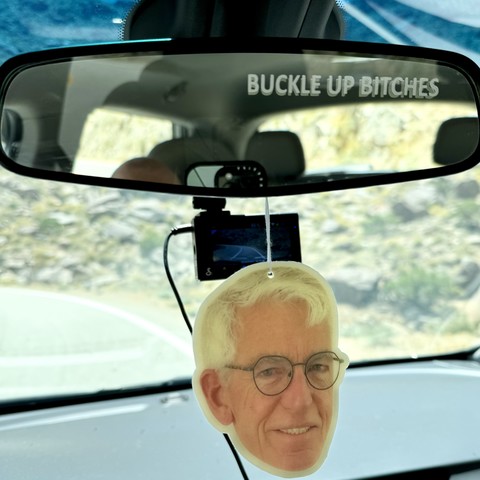 IPhone photo of my car rearview mirror with a BUCKLE UP BITCHES sticker on it & an air freshener of my husband’s head & smiling face hanging from it, beyond my windshield (& behind me) is a curvy road & a steep rocky mountainside