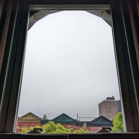 Looking through an arched window two hours after sunrise the sky is a blank slate of dense gray fog. Pointed roofs of Harlem brownstones with red brickwork are across the street, and a taller apartment building can be seen in the distance. The green leaves of trees are entering frame on the bottom left and right.