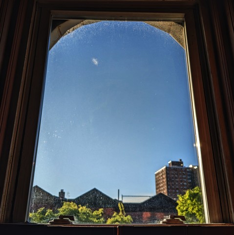 Looking through an arched window ninety minutes after sunrise the sky is cloudless and deep blue. Pointed roofs of Harlem brownstones are silhouetted across the street, and a taller apartment building can be seen in the distance. The green leaves of trees are entering frame on the bottom left and right.
