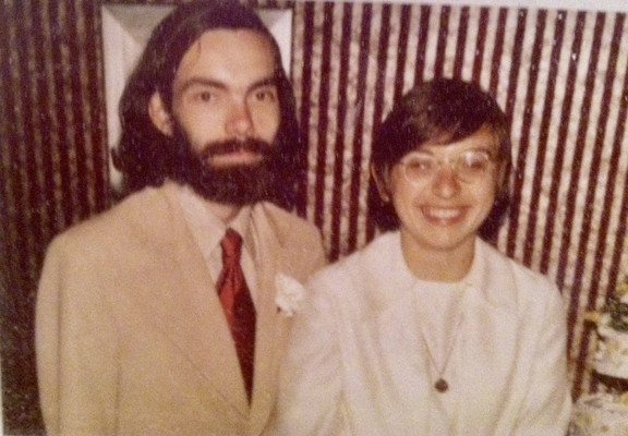 Photo of a white man with long dark hair and a beard wearing a tan suit with a red tie, standing next to a white woman with a Bob haircut and glasses wearing a white suit dress, in front of a wall with vertical stripes, taken at their wedding reception 50 years ago today. This is the only pictorial evidence of our wedding. Married by a judge wearing plaid golf pants under his robe, witnessed by our respective parents, my dad was supposed to take pictures but he’d loaded the camera wrong so ther…