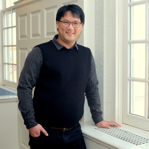 A man leaning against a window box indoors, smiling at the camera.