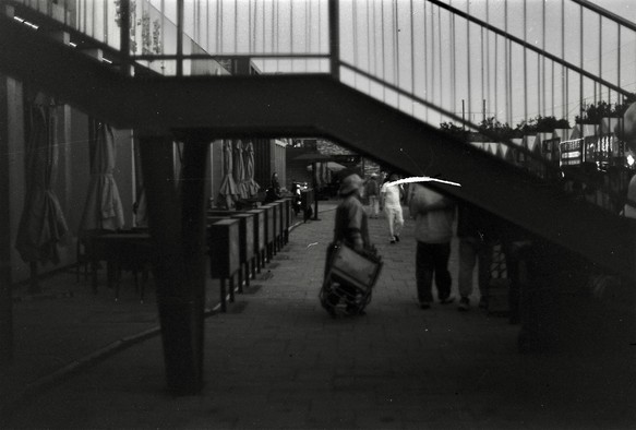 A black and white image of a person walking underneath an overpass. The image has a visible scratch from the negative.