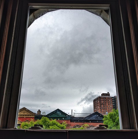 Looking through an arched window ninety minutes after sunrise the sky is full of dark gray rain clouds and there are scattered rain drops on the glass. Pointed roofs of Harlem brownstones with red brickwork are across the street, and a taller apartment building can be seen in the distance. The green tops of two trees are entering frame on the bottom and right.