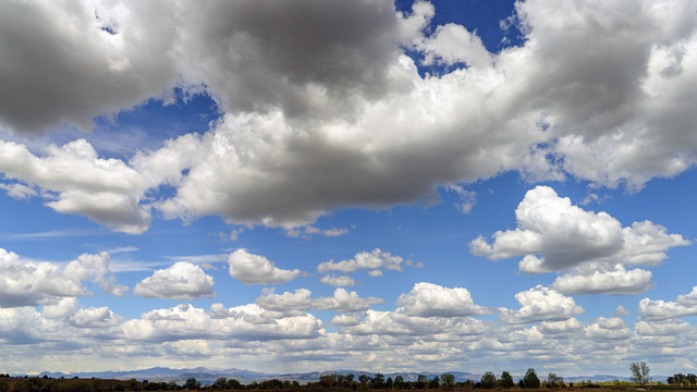 Panoramic photo of mostly white clouds in the Colorado sky. I call them Simpson Clouds, from the opening credits on The Simpsons. Except the gray bottoms of the clouds gives it a slightly sinister look, I think. The view is looking west. If you zoom in to the bottom the Mummy Range of the Rocky Mountains is the bumps at the left, and lower, but closer foothills at the center. May 2024