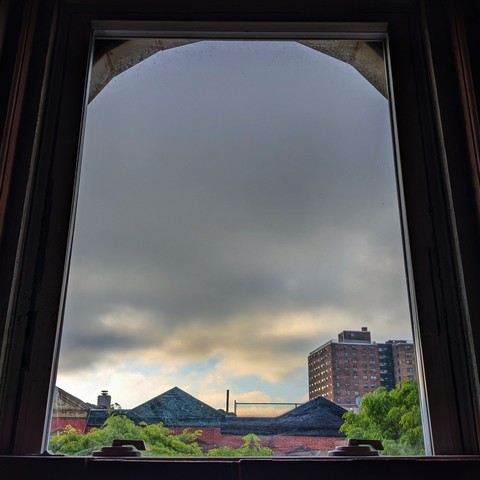 Looking through an arched window 100 minutes after sunrise the sky is full of gray storm clouds, with a patch of clearing blue sky at the horizon. Pointed roofs of Harlem brownstones with red brickwork are across the street, and a taller apartment building can be seen in the distance. The green tops of two trees are entering frame on the bottom and right.