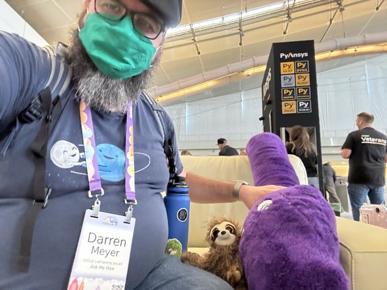 A person wearing a mask and glasses sits on a couch, holding a purple dinosaur plush toy and a small sloth plushie. Their name badge reads "Darren Meyer." People and a PyAnsys sign are visible in the background.