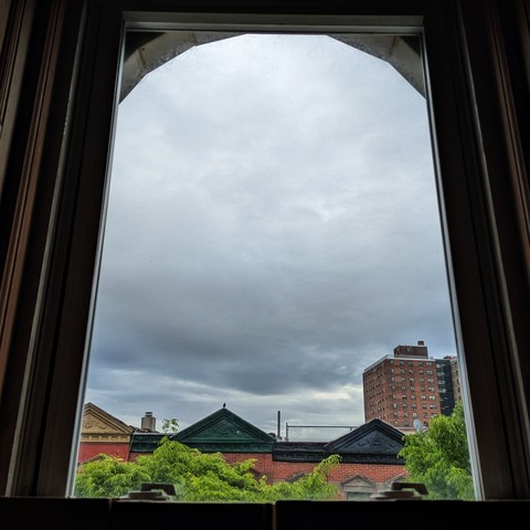 Looking through an arched window 90 minutes after sunrise the sky is full of gray storm clouds. Pointed roofs of Harlem brownstones with red brickwork are across the street, and a taller apartment building can be seen in the distance. The green tops of two trees are entering frame on the bottom and right.