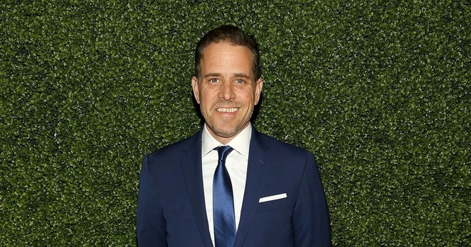 Photo of Hunter Biden standing in front of a thick hedge, wearing a navy suit and tie with a white undershirt. He’s smiling.