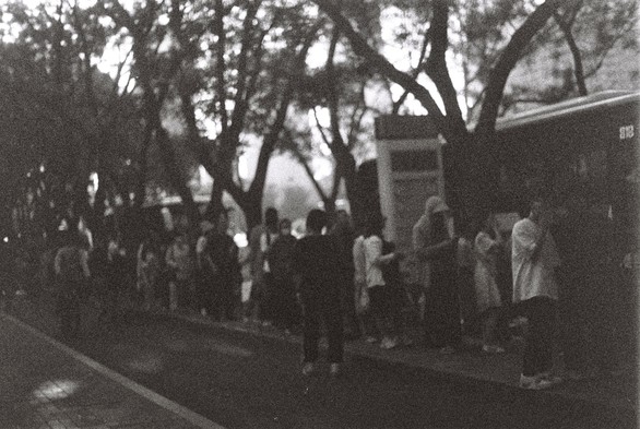 A Black and white image of people queueing for a bus in the shade.