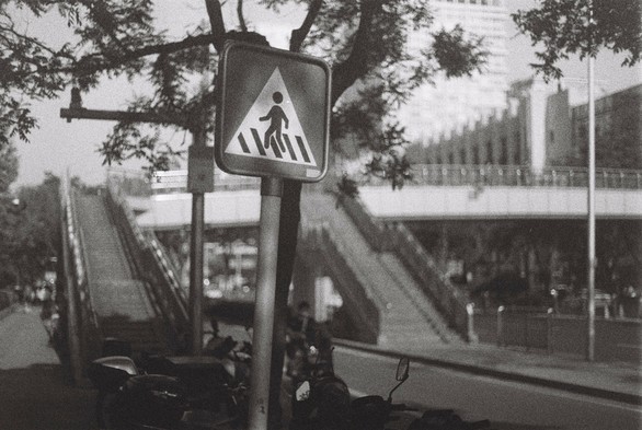 A black and white image of a road sign of pedestrian crossing set against a bridge crossing.