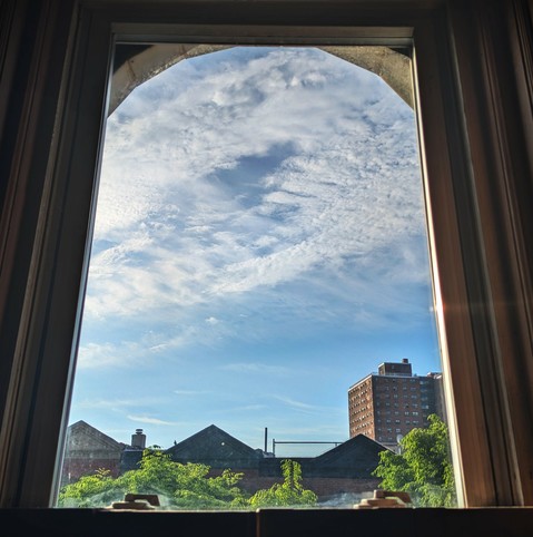 Looking through an arched window ninety minutes after sunrise the light blue sky is brushed with a large swath of crumbly white clouds. Pointed roofs of Harlem brownstones  are silhouetted across the street, and a taller apartment building can be seen in the distance. The green tops of two trees are starting to fill the frame on the bottom and right.