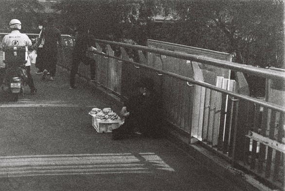 A black and white image of a man sitting on a bridge crossing the road. A person is riding an electronic bike further in front of him and some other people walking.