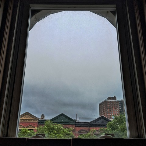 Looking through an arched window two hours after sunrise the sky is full of myrky blue-grey clouds. Pointed roofs of Harlem brownstones with red brickwork are across the street, and a taller apartment building can be seen in the distance. The green tops of two trees are starting to fill the frame on the bottom and right.