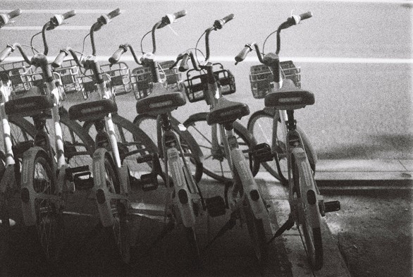 A black and white image of a line of bicycles in the shadows in front of a road.