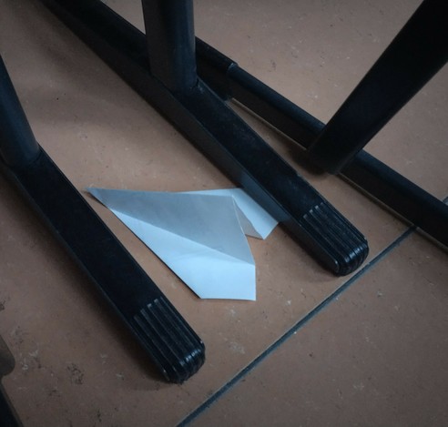 Between the metal legs of some tables and chairs rests a paper plane that crashed there, it seems, folded from a sheet of paper.
