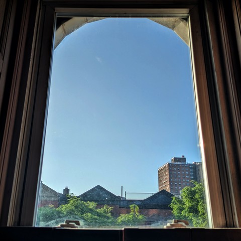 Looking through an arched window ninety minutes after sunrise the sky is blue and cloudless. Pointed roofs of Harlem brownstones are across the street, and a taller apartment building can be seen in the distance. The green tops of two trees are starting to fill the frame on the bottom.
