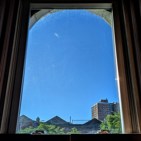 Looking through an arched window two hours after sunrise the sky is deep blue and cloudless. Pointed roofs of Harlem brownstones are silhouetted across the street, and a taller apartment building can be seen in the distance. The green tops of two trees are starting to fill the frame on the bottom and right.