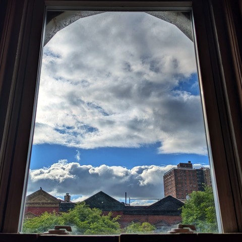 Looking through an arched window two and a half hours after sunrise the sky is deep blue and full of dramatic big puffy white clouds. Pointed roofs of Harlem brownstones with red brickwork are across the street, and a taller apartment building can be seen in the distance. The green tops of two trees are starting to fill the frame on the bottom and right.