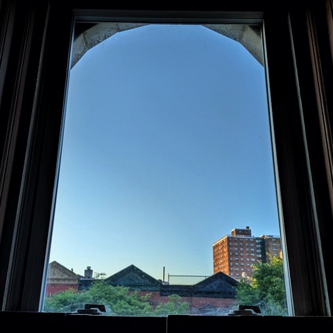 Looking through an arched window forty three minutes after sunrise the sky is deep blue and cloudless. Pointed roofs of Harlem brownstones with red brickwork are across the street, and a taller apartment building can be seen in the distance. The green tops of two trees are starting to fill the frame on the bottom and right.