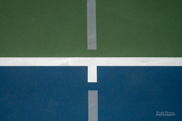 Looking down on a tennis court with a green and blue area divided horizontally by a white line an vertically by a grey pickleball line. 
