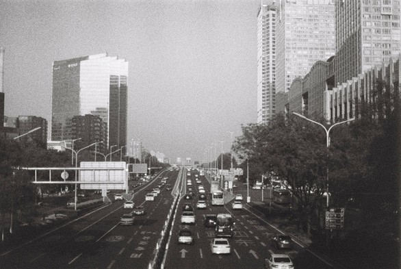 A black and white image of a semi-aerial view from an over-bridge looking over a road set against a few skyscrapers.