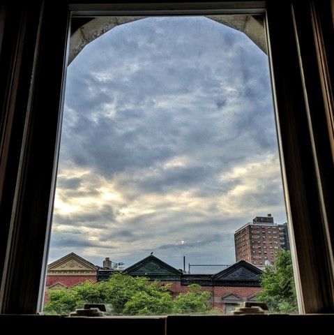 Looking through an arched window forty five minutes after sunrise the sky is full of swirling blue and parchment clouds. The sun is in the middle sky on the left making a brighter patch of cloud. Pointed roofs of Harlem brownstones with red brickwork are across the street, and a taller apartment building can be seen in the distance. The green tops of two trees are starting to fill the frame on the bottom and right.