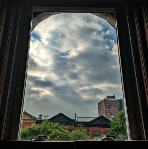 Looking through an arched window two hours after sunrise the blue sky is full of chunky white and grey clouds. Pointed roofs of Harlem brownstones with red brickwork are across the street, and a taller apartment building can be seen in the distance. The green tops of two trees are starting to fill the frame on the bottom and right.