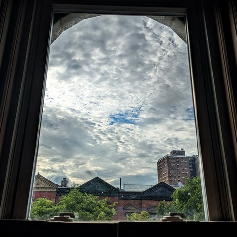 Looking through an arched window two hours after sunrise the sky is full of complex white and grey clouds with a single small patch of blue sky visible in the center. Pointed roofs of Harlem brownstones with red brickwork are across the street, and a taller apartment building can be seen in the distance. The green tops of two trees are starting to fill the frame on the bottom and right.