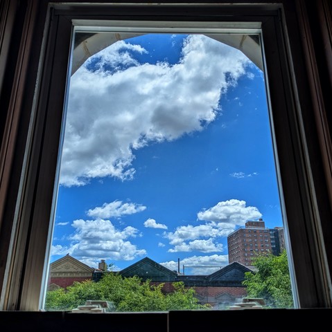 Looking through an arched window just before midday the deep blue sky is full of big fluffy white clouds. Pointed roofs of Harlem brownstones with red brickwork are across the street, and a taller apartment building can be seen in the distance. The green tops of two trees are starting to fill the frame on the bottom and right.
