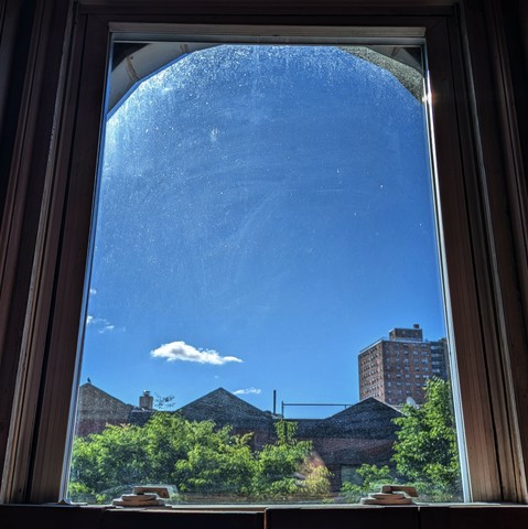 Looking through an arched window three hours after sunrise the deep blue sky has one small white cloud above the horizon. Pointed roofs of Harlem brownstones with red brickwork are silhouetted across the street, and a taller apartment building can be seen in the distance. The green tops of two trees are starting to fill the frame on the bottom and right.
